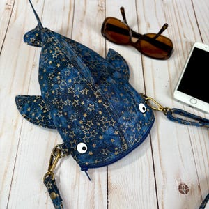 May include: A blue fish-shaped crossbody bag with a gold star pattern. The bag has a zipper closure, white googly eyes, and a detachable strap. Sunglasses and a smartphone are next to the bag.