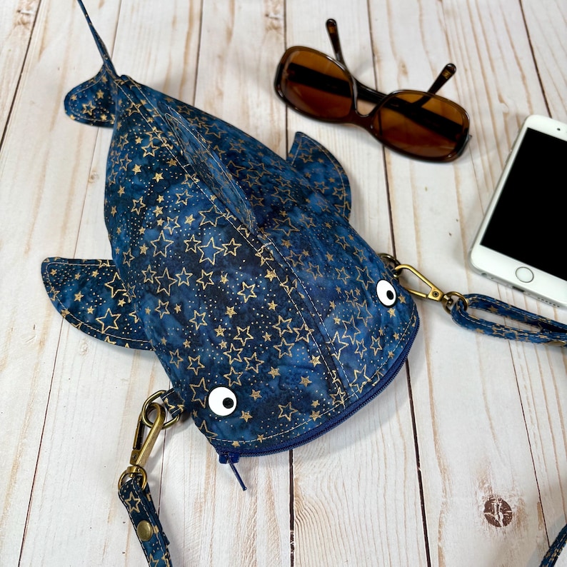 May include: A blue fish-shaped crossbody bag with a gold star pattern. The bag has a zip closure, white googly eyes, and a detachable strap. Sunglasses and a smartphone are next to the bag.