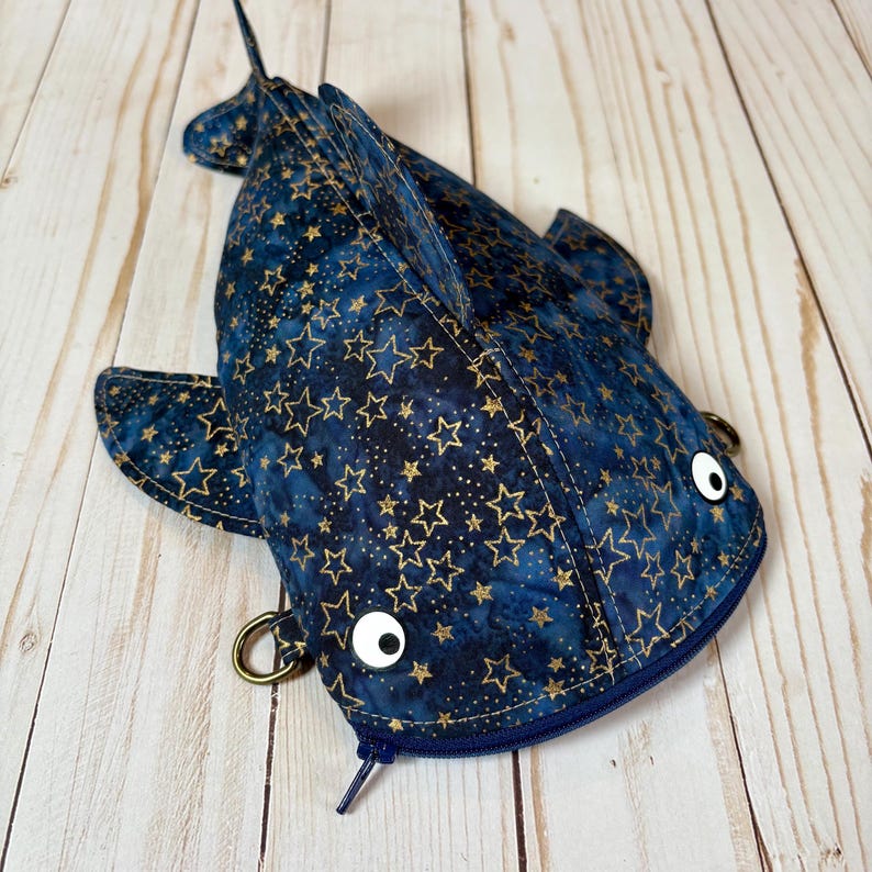 May include: A navy blue fish-shaped pouch with a gold star pattern. The pouch has a zip closure, two white button eyes, and metal ring attachments. The design includes fins and a tail. The pouch is a fun, handmade accessory, perfect for carrying small items.