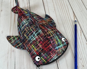 May include: A shark-shaped pencil case featuring a black fabric base with a multi-colored crosshatch pattern of red, yellow, blue, and green lines. It has two white circular eyes and a black zipper closure. A blue pencil is placed next to it.