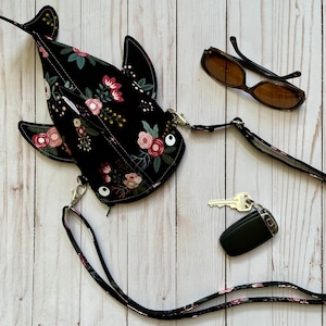 May include: A black fabric purse shaped like a shark with floral print and a black adjustable strap. The purse is on a white wooden surface with sunglasses and keys.