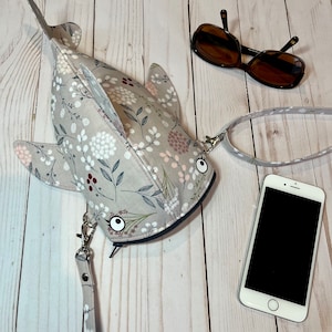 May include: A gray and white floral print fabric handbag shaped like a shark with a zipper closure and a long strap. The bag has a white background with a floral pattern in shades of gray, white, and pink. The bag has two large white eyes.