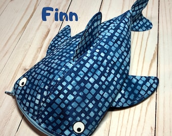 May include: A whale shark-shaped fabric pencil case featuring a dark blue and light blue square grid pattern. The case has two white circular eyes with black pupils, a dorsal fin, side fins, a tail, and a zipper closure. The name Finn is printed in blue.