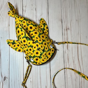 May include: A yellow and black fabric bag shaped like a fish with a sunflower pattern. The bag has a long strap and a zipper closure.