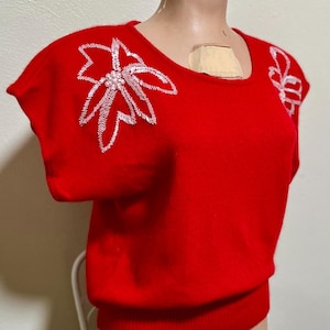 May include: A red knitted sweater with white embroidered flowers on the shoulders. The sweater has short sleeves and a ribbed hem.