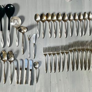 May include: A collection of vintage silverware, including forks, spoons, knives, and serving utensils. The silverware has a floral design on the handles and is arranged on a light gray surface. The set includes various sizes of spoons and forks.