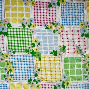 May include: Close-up of a quilted fabric with a patchwork design. The squares are in various colors, including yellow, green, blue, and pink, with a gingham pattern. Floral accents in pastel colors are scattered throughout the design.
