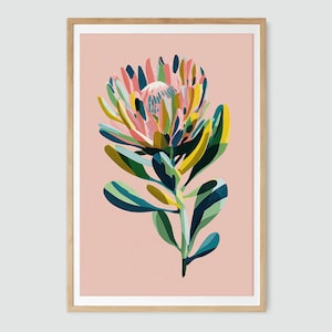 Puede incluir: Una impresión artística enmarcada con una colorida ilustración de una flor de protea. La flor tiene pétalos y hojas rosas, amarillos, azules y verdes sobre un fondo rosa suave. La obra de arte está en un marco de madera.