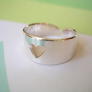 May include: A silver-colored ring with a heart-shaped cutout. The ring has a thick band and an open design. The heart is cut out of the band, creating a negative space. The ring is on a white surface with a green and white background.