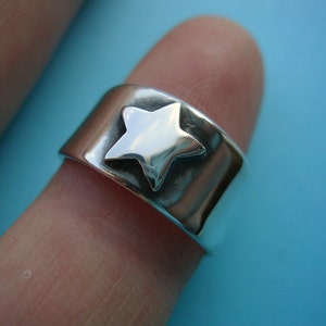 May include: A wide, silver-colored ring with a raised star design. The star is centered on the band and has a polished finish. The ring is shown on a finger against a light blue background.