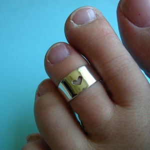 May include: A silver toe ring with a heart-shaped cutout. The ring is wide and wraps around the toe. The background is a solid light blue.