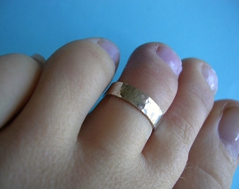 May include: A hammered gold-colored toe ring. The ring is a simple band with a textured surface, worn on a toe. The background is a solid light blue.