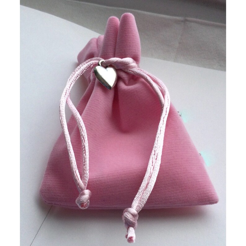 May include: A small, pink velvet drawstring bag with a silver heart charm. The bag is closed with a matching pink cord. The bag is likely used for jewelry or small gifts.