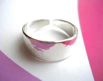 May include: A hammered silver ring with an open design. The ring has a textured surface and a bright, reflective finish. The background features a white and pink color scheme, creating a visually appealing contrast.
