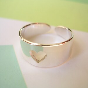 May include: A silver-colored ring with a heart-shaped cutout. The ring is a wide band with an open design, allowing for slight size adjustment. The heart is centered on the band, adding a touch of romance to the jewelry.