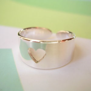 May include: A silver-colored ring with a heart-shaped cutout. The ring has a wide band and an open design. The heart is centered on the band. The ring is likely made of sterling silver.