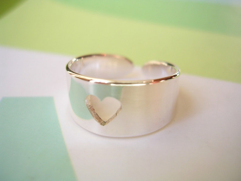 May include: A silver-colored ring with a heart-shaped cutout. The ring has a wide band and an open design. The heart is centered on the band. The ring is likely made of sterling silver.