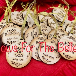 May include: A collection of wooden ornaments with religious text and scripture verses. The ornaments are round and have a rustic, wood-burned look. The ornaments are decorated with a simple leaf design. The text on the ornaments includes "Good Shepherd", "Lamb of God", "Savior", "Messiah", "The Word", and "Redeemer".