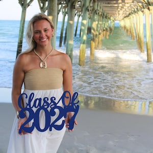 May include: A dark blue cutout sign featuring the text Class Of 2027 in a cursive script font. The sign is held in front of a white and tan strapless dress. The accessory is made of a solid, flat material with a smooth, matte finish.