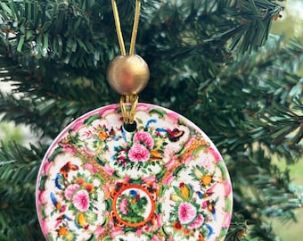May include: A round, flat ceramic ornament featuring a colorful floral and butterfly pattern in shades of pink, green, and orange. The ornament is suspended by a gold-colored string with a single metallic gold bead at the top for hanging.