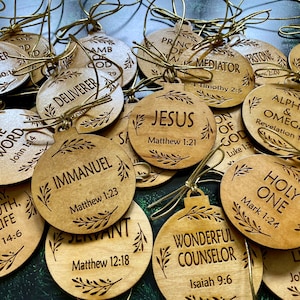 May include: A collection of wooden Christmas ornaments with gold string hangers. Each ornament is round and features a different religious word or phrase, such as "Jesus" and "Immanuel", along with a corresponding Bible verse.