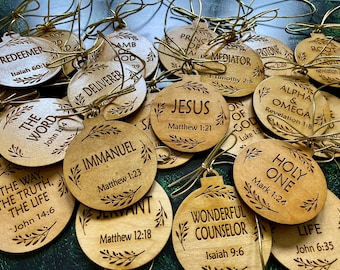 May include: A collection of wooden Christmas ornaments with gold string hangers. Each ornament is round and features a different religious word or phrase, such as "Jesus" and "Immanuel", along with a corresponding Bible verse.
