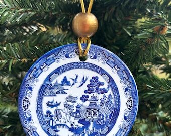 May include: A round ceramic ornament featuring a blue and white willow pattern design. The ornament is suspended by a gold cord with a single gold bead at the top. The intricate illustration depicts pagodas, trees, and birds in a classic style.