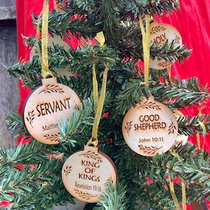 May include: Wooden Christmas ornaments with religious themes. The ornaments are round and have a rustic brown color. The ornaments feature the words "Servant", "Good Shepherd", "King of Kings", and "Holy".