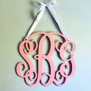 May include: A pink wooden monogram with the letters "S B C" in a decorative script font. The monogram is hanging from a white ribbon.
