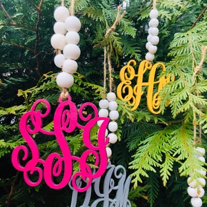 May include: Three wooden ornaments with personalized monograms. The ornaments are painted in pink, gray, and yellow. Each ornament is strung with a white beaded garland.