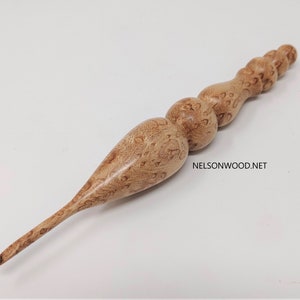 May include: A wooden crochet hook with a natural wood finish and a textured surface. The hook has a long, thin handle with a rounded tip. The handle is decorated with multiple rounded bumps.