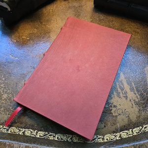 May include: A rectangular notebook with a textured, deep red cover and a matching red ribbon bookmark extending from the bottom left corner. The spine features visible raised bands, and the cover has a consistent, fine-grained surface texture.