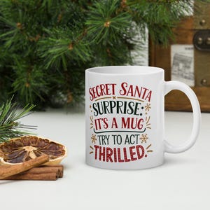 Può includere: Una tazza in ceramica bianca con un design rosso e nero che dice "Secret Santa Surprise: It's a Mug Try to Act Thrilled."