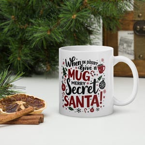 Può includere: Tazza in ceramica bianca con un design rosso e nero. La tazza dice "When in doubt give a mug merry Secret Santa!" con fiocchi di neve e bastoncini di zucchero intorno al testo.