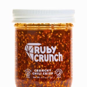 May include: A clear glass jar filled with a red chili crisp. The jar has a white lid and a label with the text "RUBY CRUNCH" and "CRUNCHY CHILI CRISP". The label also indicates the product is gluten-free and has a mild spice level. Net weight is 6.2 oz (175g).