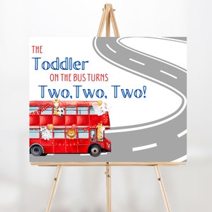 Op de afbeelding: Een verjaardagsbord met een rode dubbeldekkerbus, dieren en de tekst "The Toddler on the Bus Turns Two, Two, Two!". Een grijze weg loopt over de rechterbovenhoek. Het bord staat op een houten ezel.