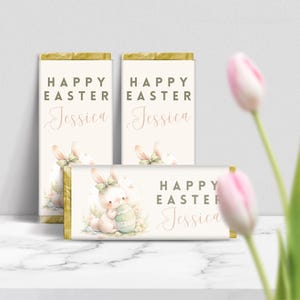 Puede incluir: Tres envoltorios de barra de chocolate blanco con un conejito rosa sosteniendo un huevo de Pascua. Los envoltorios tienen un borde dorado y el texto "Feliz Pascua Jessica" en ellos.