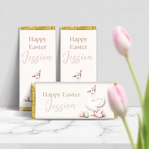 May include: Three white chocolate bar wrappers with a gold border. Each wrapper features a watercolor illustration of a goose wearing a pink hat and holding pink Easter eggs. The text "Happy Easter Jessica" is printed on each wrapper.
