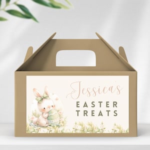 Może przedstawiać: Brązowe pudełko kartonowe z uchwytem, ozdobione naklejką z akwarelową ilustracją królika trzymającego jajko wielkanocne. Naklejka zawiera również tekst "Jessica's Easter Treats".