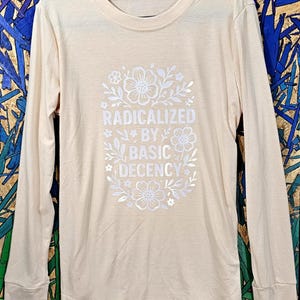 May include: A long-sleeved, pale peach-coloured shirt featuring the white text "RADICALIZED BY BASIC DECENCY" within a floral design. The shirt hangs on a black hanger, set against a vibrant, multi-coloured backdrop.