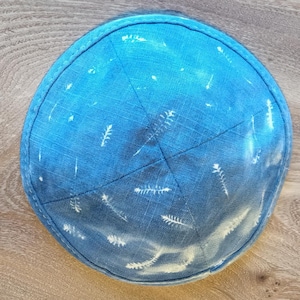 Pode incluir: Uma tigela decorativa redonda azul com um efeito gradiente, passando de um azul mais escuro no topo para um tom mais claro na parte inferior. Padrões brancos em forma de pena estão espalhados pela superfície. A tigela está sobre uma superfície de madeira.