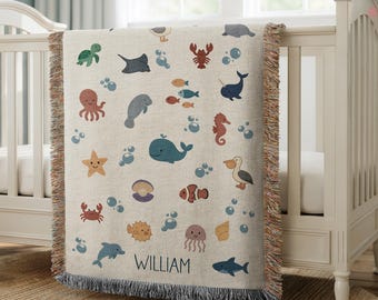 May include: A beige baby blanket with a sea life pattern, including whales, fish, and seahorses. The blanket has a fringed edge and the name "WILLIAM" is printed at the bottom. The blanket is draped over a white crib.