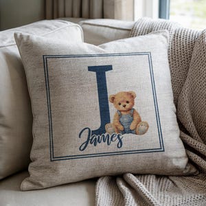 May include: A beige square pillow with a navy blue border and the letter "J". A teddy bear wearing blue overalls sits next to the letter. The name "James" is written in cursive below the letter.