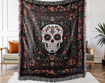 May include: Black woven throw blanket featuring a Day of the Dead sugar skull design. The skull is white with floral accents and blue and orange flower eyes. The blanket has a floral border and fringe.