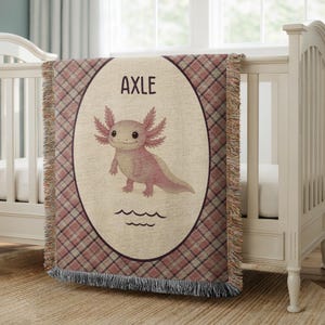 May include: A baby blanket with a plaid pattern in shades of pink and brown. The center features an oval design with an axolotl illustration and the name "AXLE" in black letters. The blanket has a fringed edge.