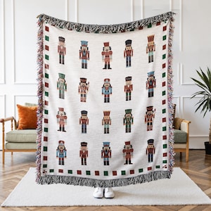 May include: A cream-colored woven throw blanket featuring a repeating pattern of colorful nutcracker soldiers. The blanket has a border of green and red squares and a fringed edge. The nutcrackers are in various uniforms.
