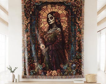 May include: A woven tapestry featuring a Day of the Dead themed design. The artwork depicts a female skeleton figure in a stained-glass style, surrounded by floral elements in shades of brown, orange, green, and red. The tapestry has a fringed edge.