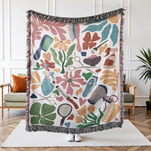 May include: A white woven throw blanket with a colorful pattern of salon tools and floral designs. The design includes combs, scissors, hair dryers, and flowers in shades of pink, blue, green, and orange. The blanket has a black fringe.