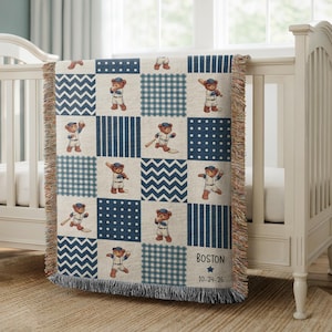 May include: A baby blanket with a patchwork design featuring baseball-playing teddy bears in a navy blue, white, and beige color scheme. The blanket has a fringed edge and includes the name "Boston" and the date "10-24-26".