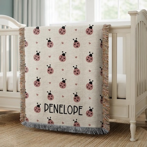 May include: Beige baby blanket with a repeating pattern of pink ladybugs and small pink hearts. The name "PENELOPE" is printed in black script. The blanket has a fringed edge and is draped over a white crib.
